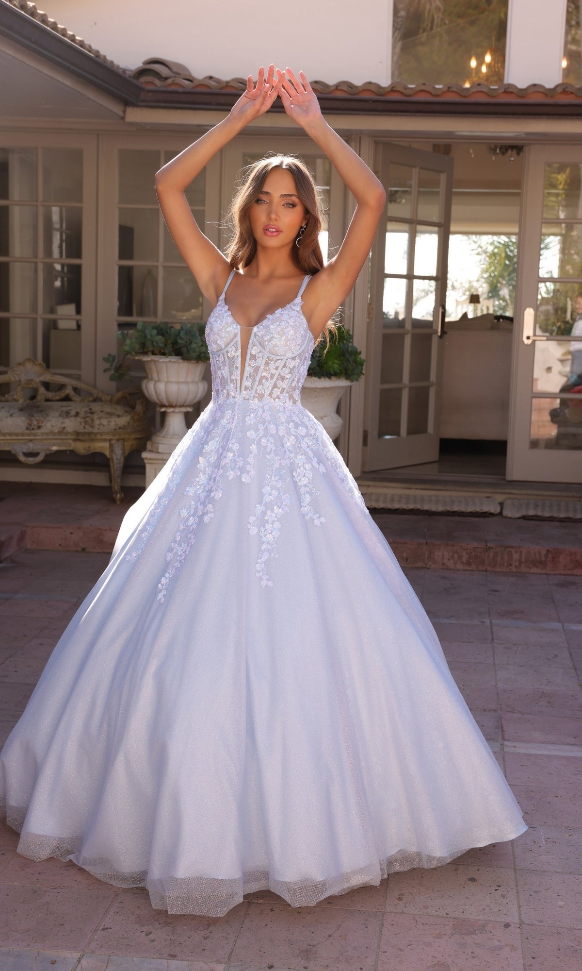 A woman wearing the Long Corset Glitter Prom Ball Gown H1357, a white floor-length dress with floral lace details, stands with her arms raised in front of a house.